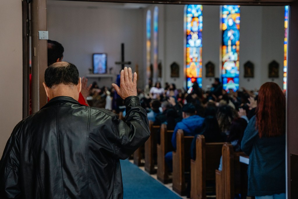 A man in a leather jacket lifts his hand in prayer inside a church.