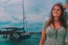 Kelsey Granger smiles while leaning against a mast with wind-blown hair. Behind her, a sailboat floats.