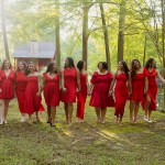 Twelve members of Alpha Pi Omega sorority, all wearing red dresses, walk hand in hand through a wooded area near a cabin.