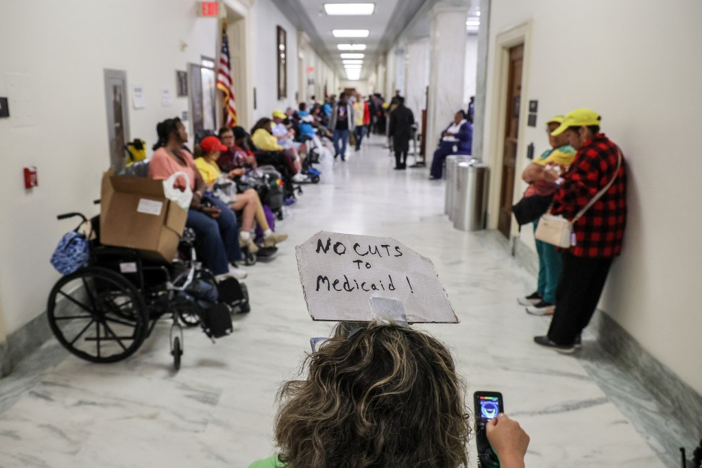 A person using a wheelchair wears a "no cuts other than Medicaid" placard on their head.