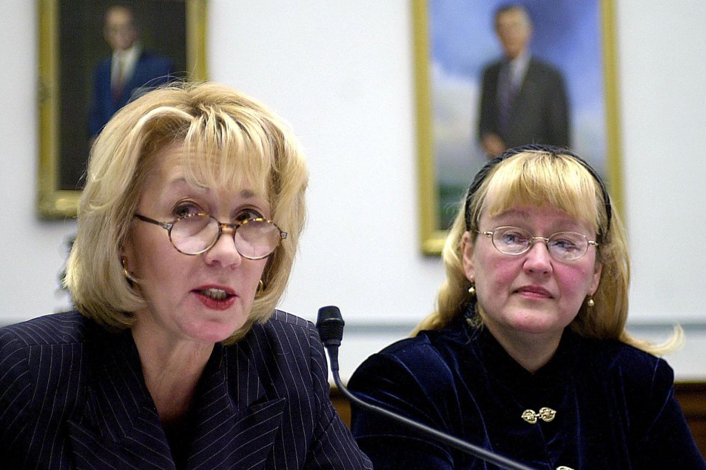 Two women sit at a congressional hearing; one speaks into a microphone while the other listens, both wear glasses and formal attire.