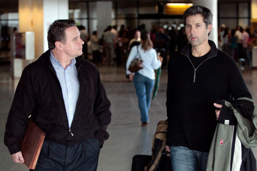 David Goldman, wearing a black sweater, walks through a busy airport terminal alongside Rep. Chris Smith, who is holding a briefcase and looking at Goldman. Travelers are visible in the background.