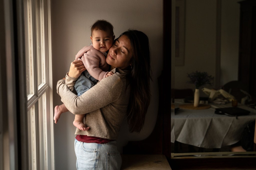 Jewel Lazaro holds her baby Selah close near a window in their apartment. Jewel’s eyes are closed as she hugs Selah, who looks out with a calm expression.
