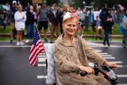 An older woman in a rain poncho rides a mobility scooter, smiling and holding an American flag during a protest. People with signs and flags are visible in the blurred background.