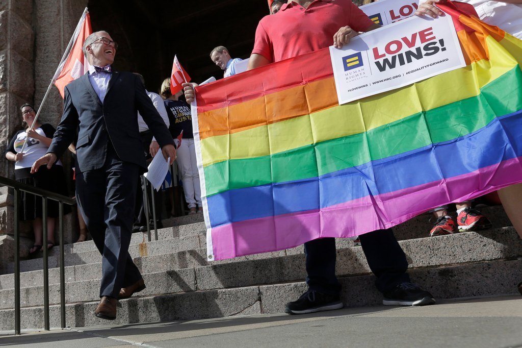 Jim walks down steps laughing beside a rainbow flag and a sign that says love wins".