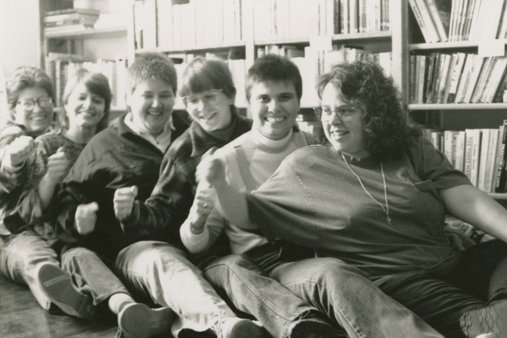 A row of people lean into each other while seating on the floor in front of stacks of books.