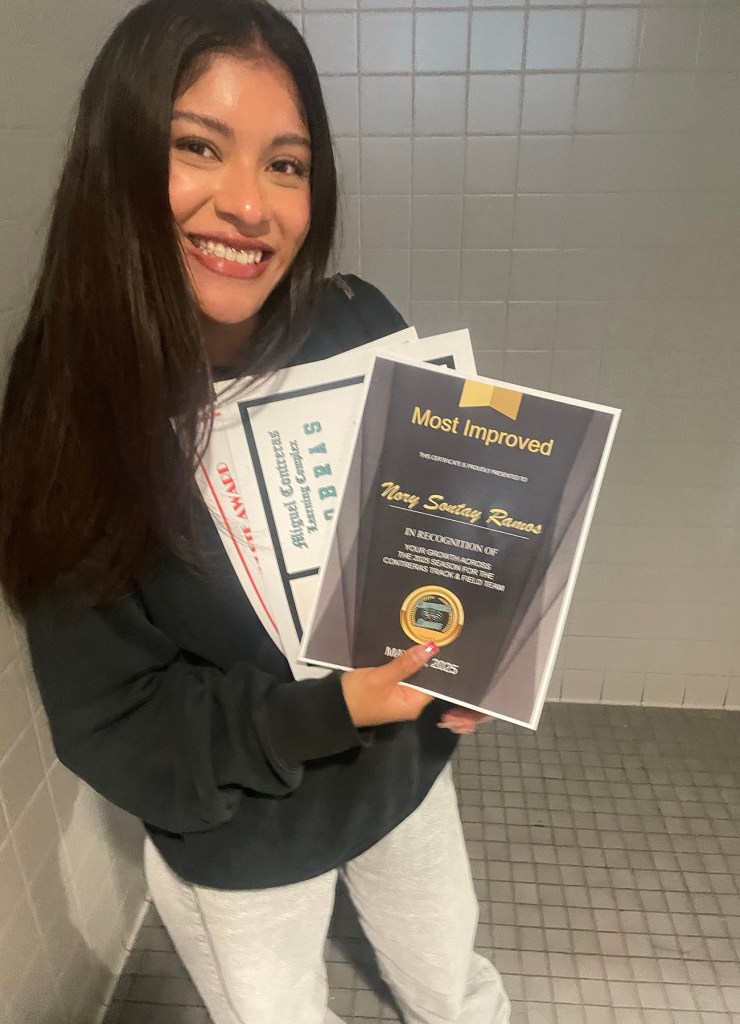 Nory Sontay Ramos holds a stack of school certificates, including one labeled “Most Improved,” and smiles for the camera.