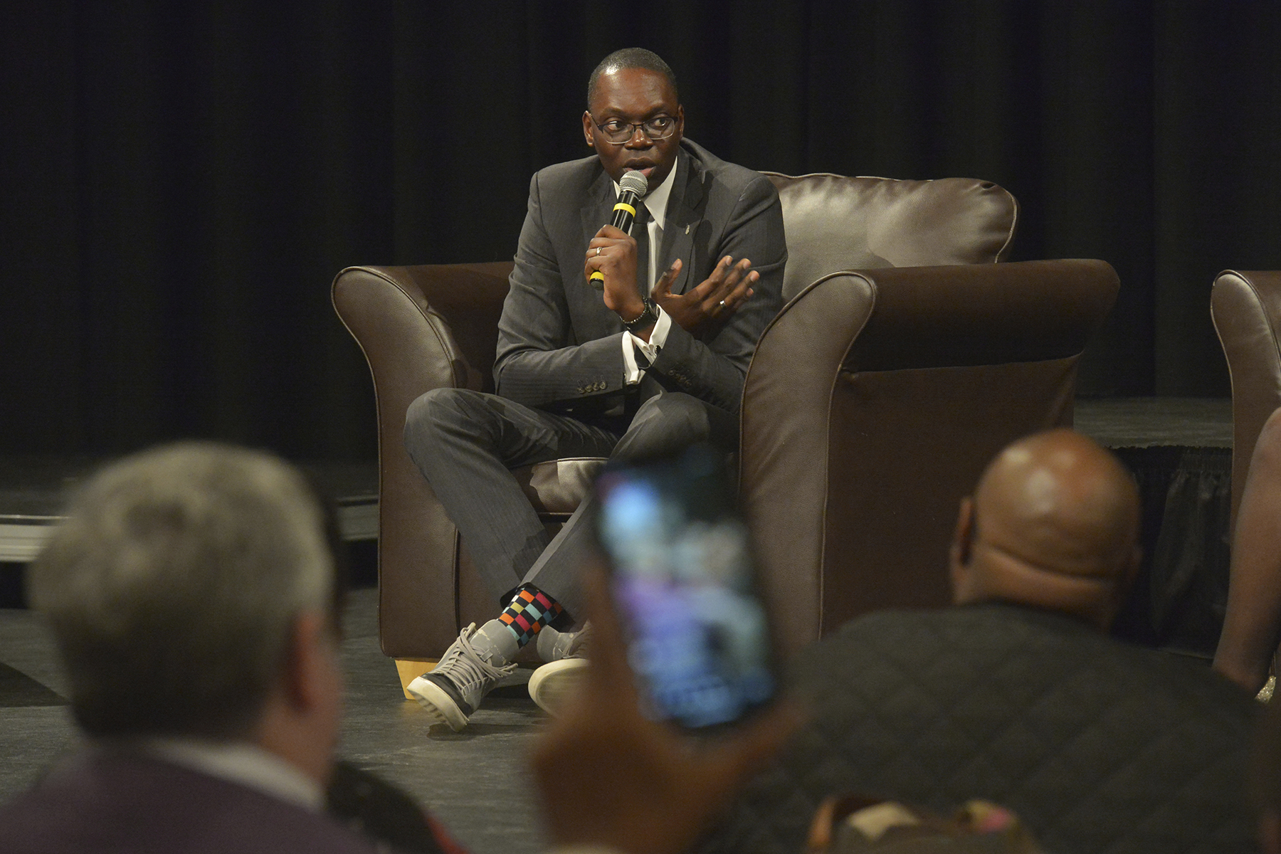 Garlin Gilchrist sits onstage in a suit and sneakers, speaking to a crowd.