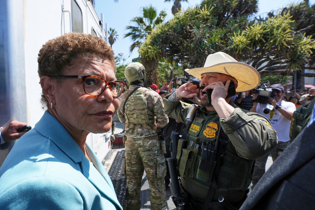 Karen Bass stands in front of a Border Patrol agent who is on the phone.