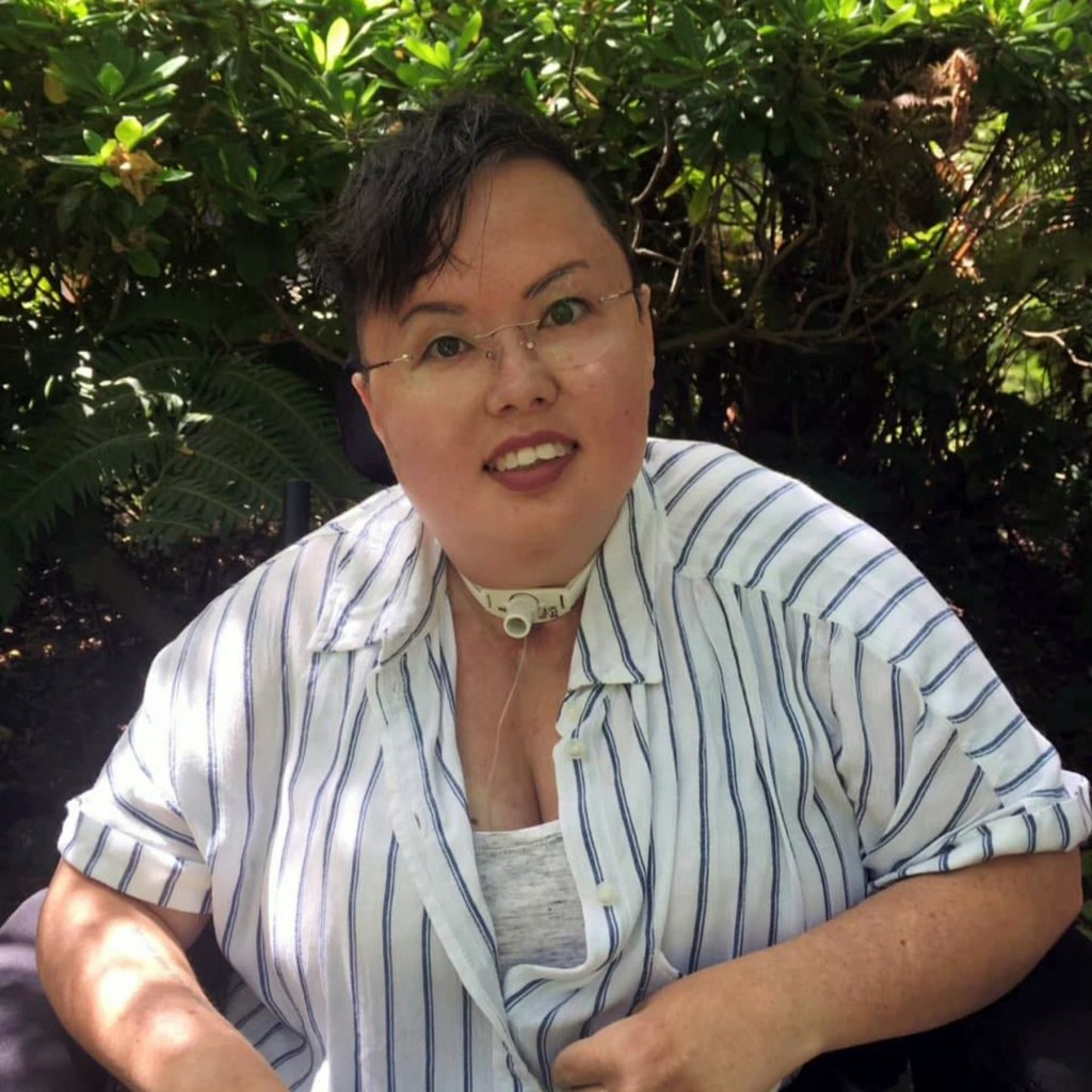 Portrait of Stacey Park Milbern outdoors, wearing glasses and a striped shirt, smiling at the camera. She has a tracheostomy tube visible at her neck.