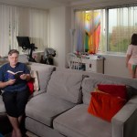 Brian reads on a couch as his daughter looks out the window behind him in their living room.