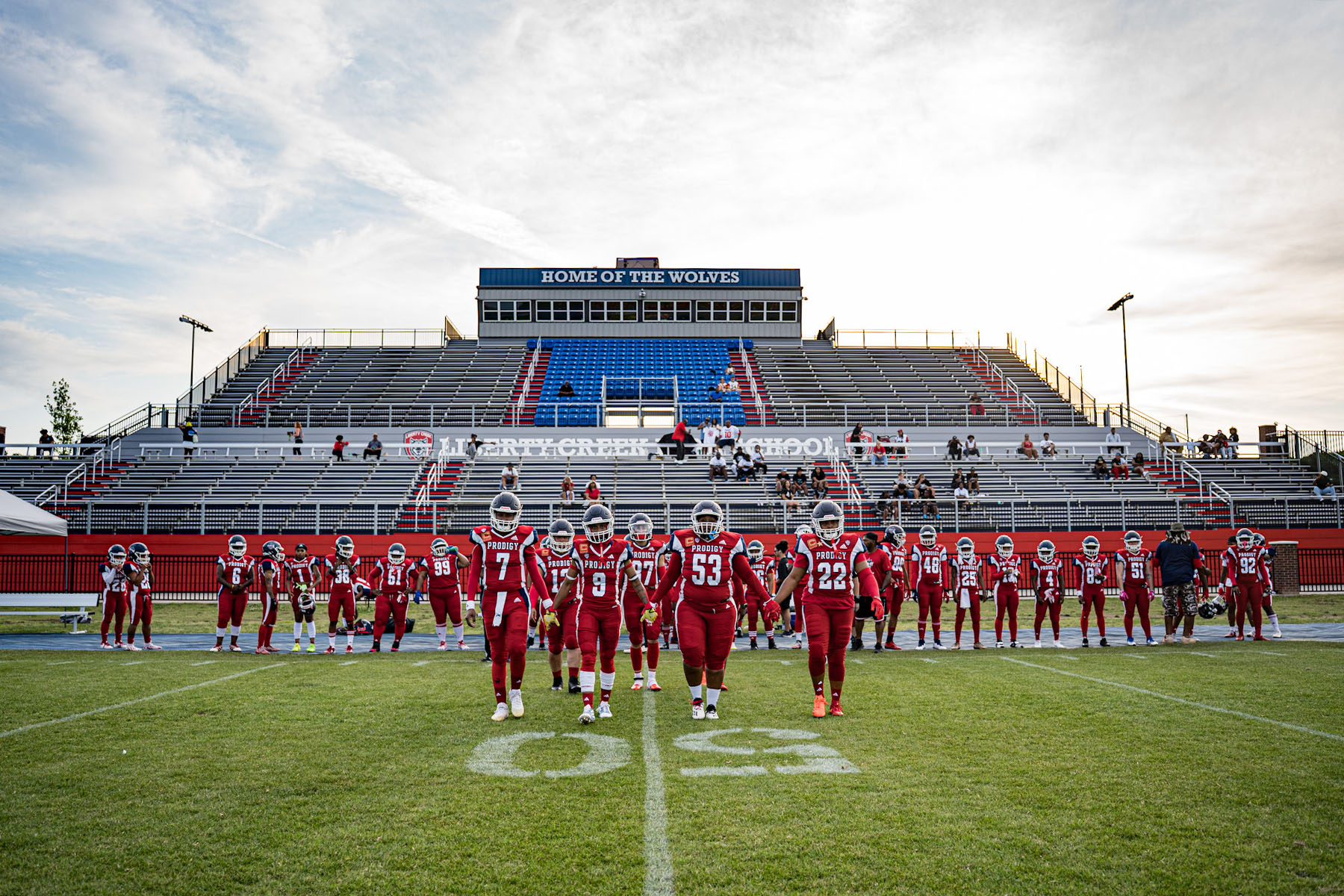 A women’s tackle football team, the Washington Prodigy, walks hand in hand across the field in red uniforms, with rows of teammates standing behind them and empty stadium bleachers in the background.