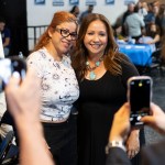 Two women smile for photos being taken by two cell phones.