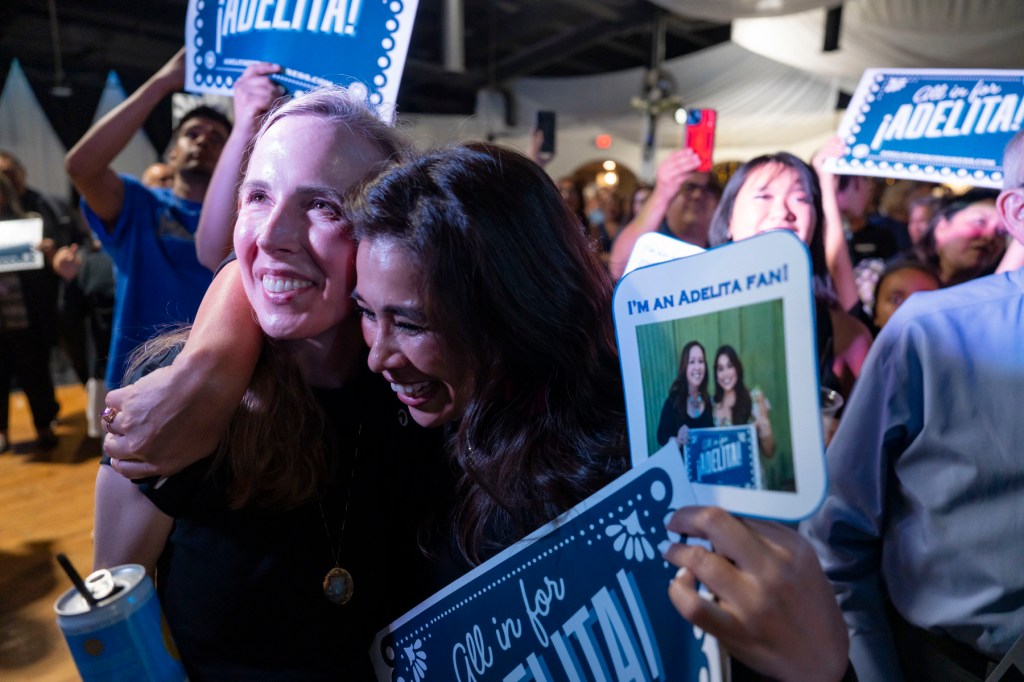 A crowd of smiling and hugging supporters hold fans that say "All in for Adelita!"