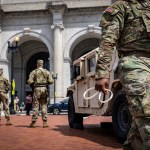 A National Guard walks while holding zip ties past a military vehicle as pedestrians are in the background.