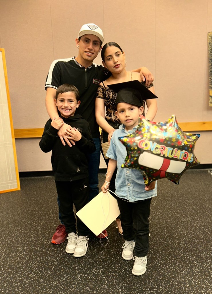 A family of four poses indoors at a celebration. The parents stand behind their two young sons, one wearing a graduation cap and holding a balloon.