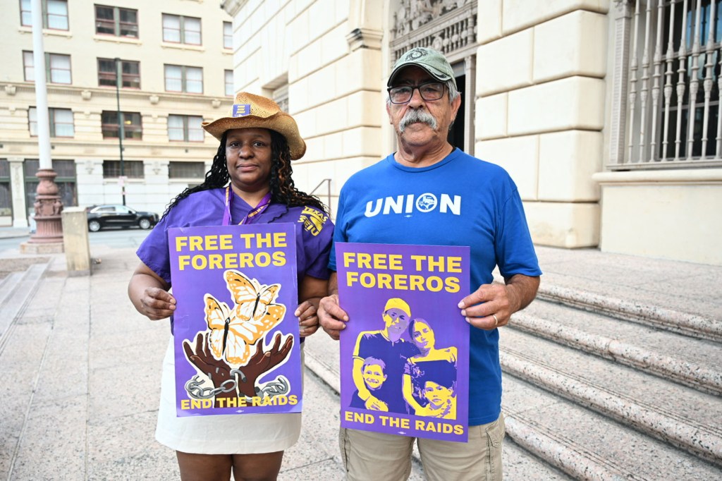 Two demonstrators hold purple posters reading “Free the Foreros, End the Raids,” with stylized portraits of the family alongside butterfly and broken chain symbols.