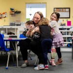 Toddlers give their teacher a hug in a classroom.