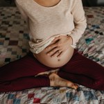 A pregnant person holds their belly while sitting cross-legged on a bed.