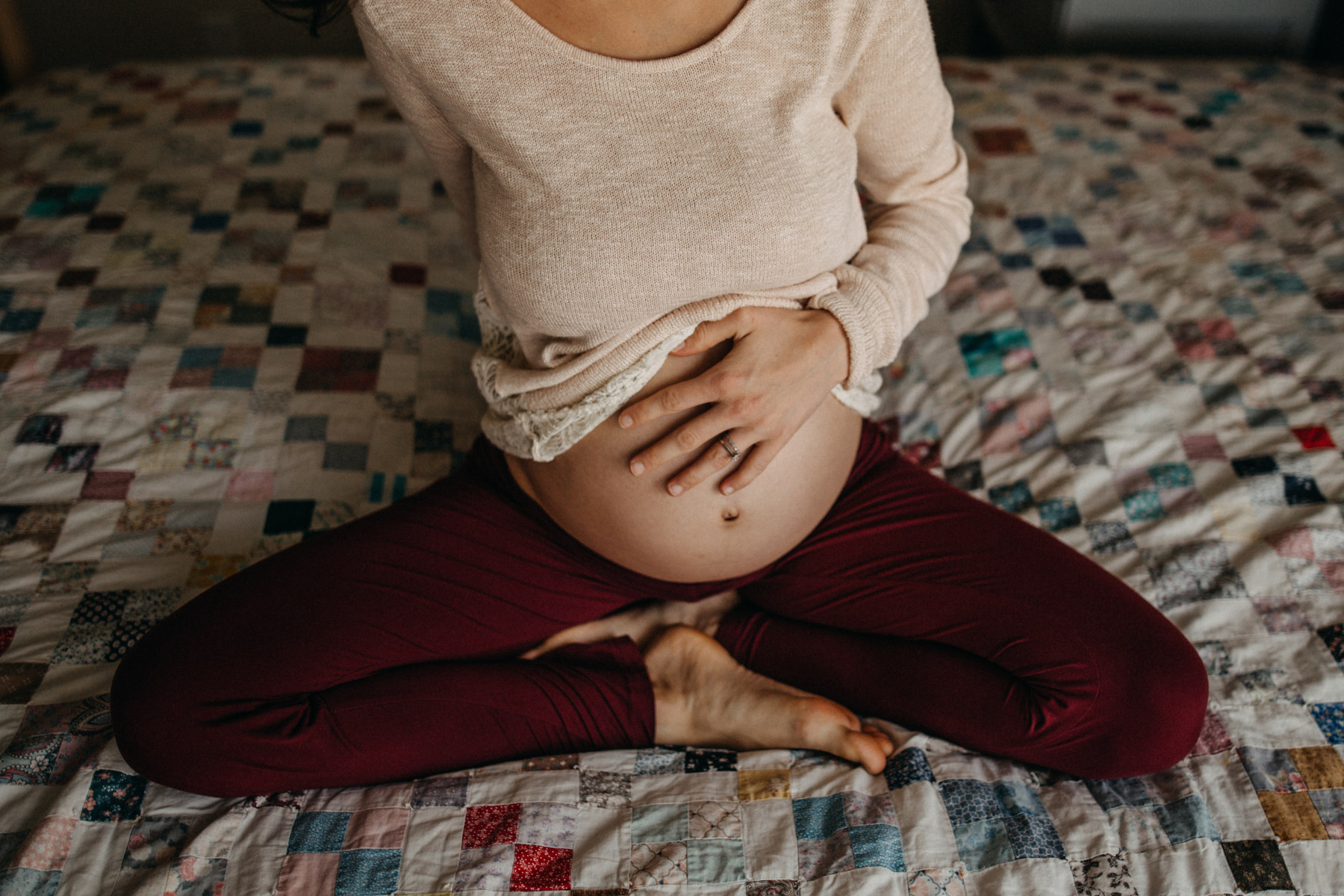 A pregnant person holds their belly while sitting cross-legged on a bed.