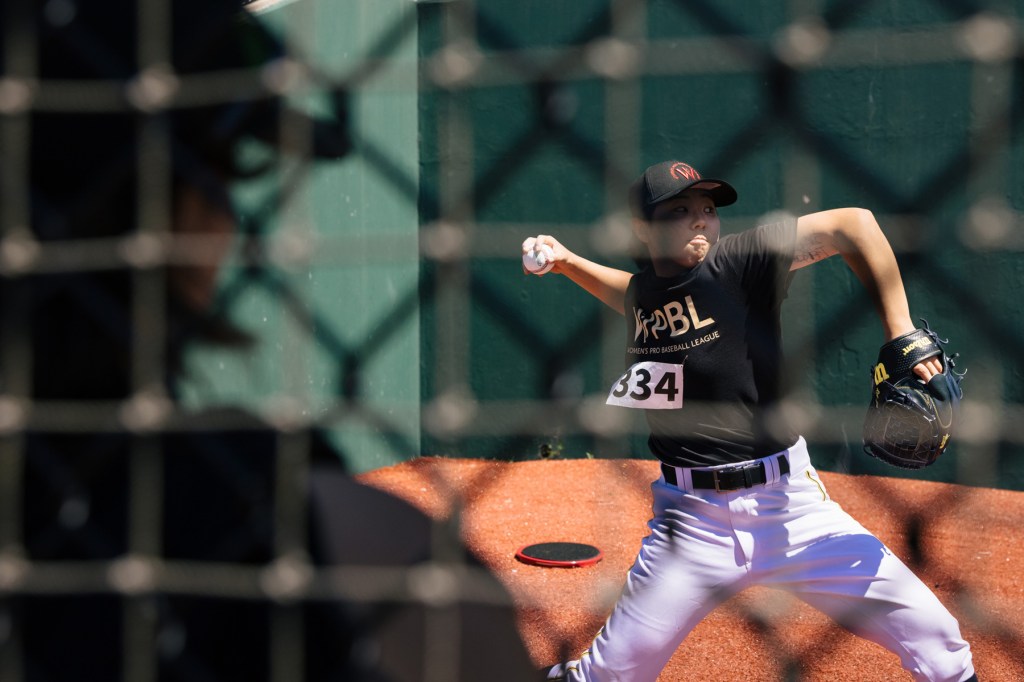 A female baseball player throws a fast pitch seen through a fence.