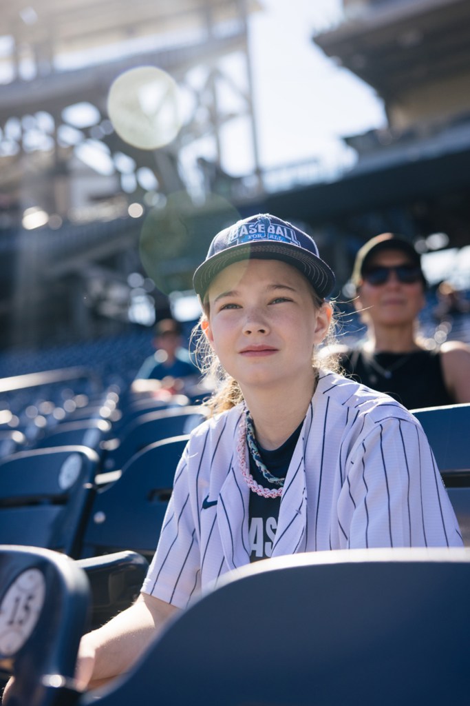 A young girl sits in the stands wearing a baseball jersey.