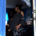 A pregnant woman stands inside a small blue-painted home, one hand on her belly, looking away from the camera toward the dimly lit room behind her.