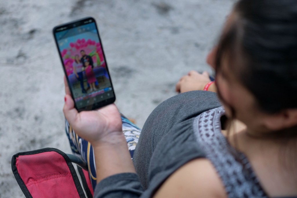 A pregnant woman sits in a red folding chair, looking down at her smartphone showing a photo of her family against a pink backdrop.