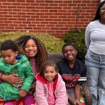 A woman smiles into the camera while holding one young child as three other children sit and stand beside her smiling.