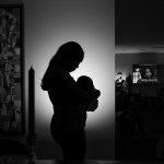 A Black mother holds an infant in silhouette against a candlelit homescape.