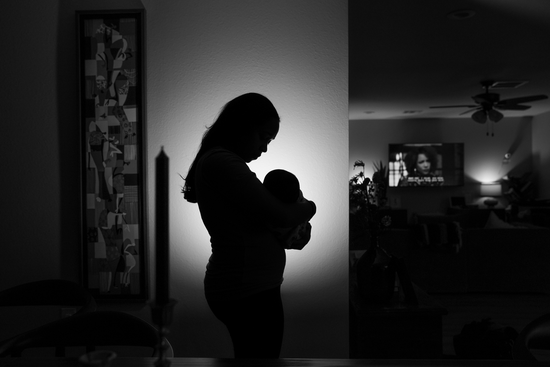 A Black mother holds an infant in silhouette against a candlelit homescape.