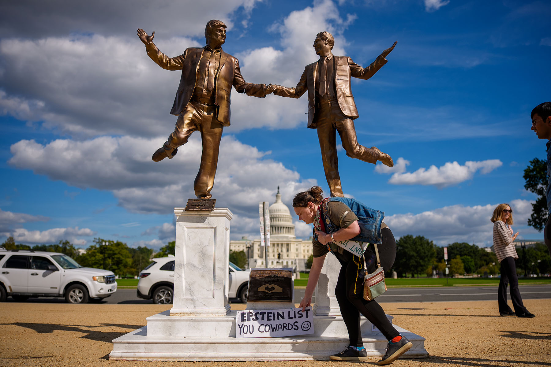 A woman picks up a sign that reads "Epstein List You Cowards" at the base of a statue depicting U.S. President Donald Trump and Jeffrey Epstein holding hands on the National Mall.