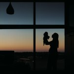 A woman holds a newborn baby up in the air, silhouetted against a window and sunset background.