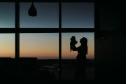 A woman holds a newborn baby up in the air, silhouetted against a window and sunset background.