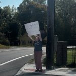 A woman holds a sign on a street corner with a truck parked alongside her.