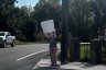 A woman holds a sign on a street corner with a truck parked alongside her.