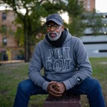 A man in a gray hoodie sits on a park bench, hands clasped.
