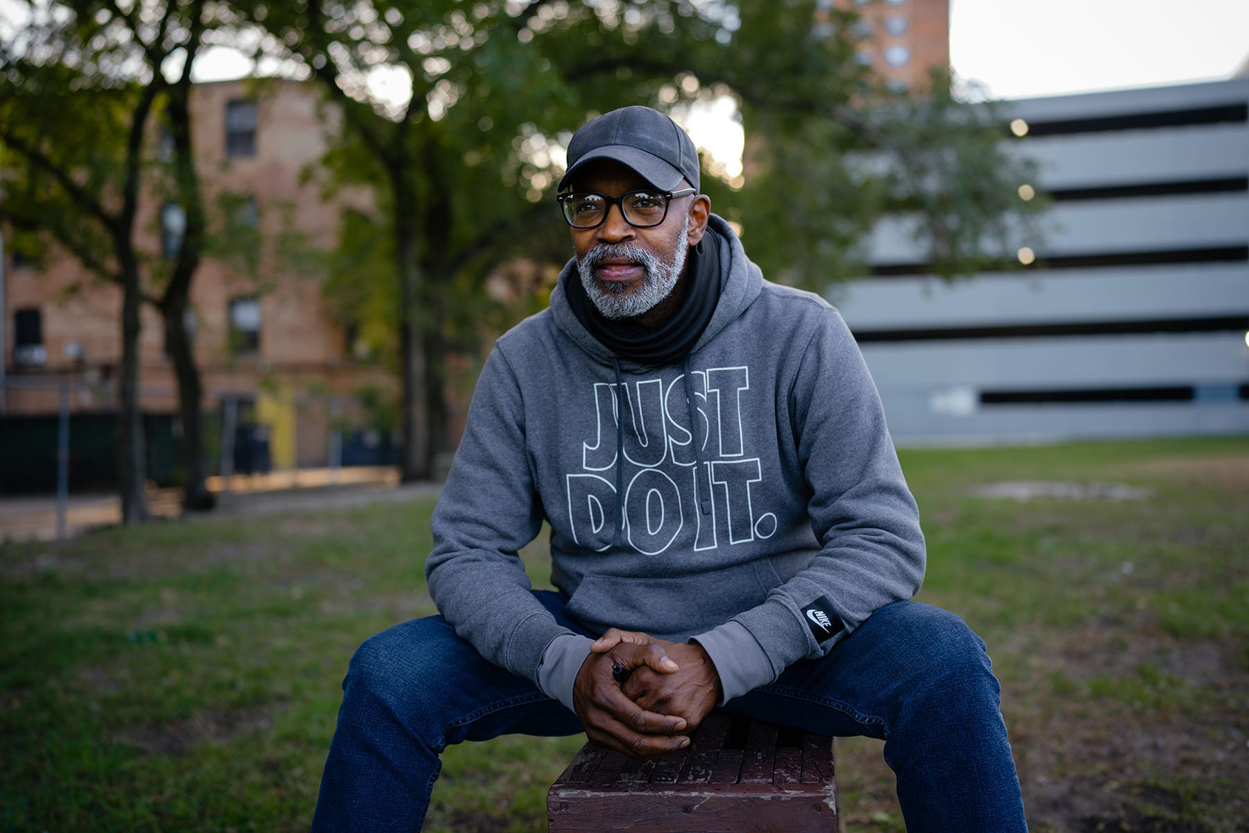 A man in a gray hoodie sits on a park bench, hands clasped.