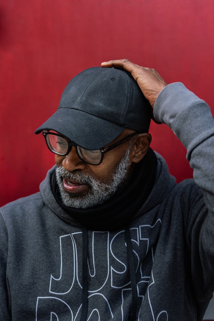 A man in a gray hoodie and black cap stands against a red wall, looking down, on Chicago’s North Side.
