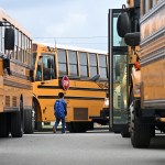A young child wearing a blue backpack walks between a row of yellow school buses.