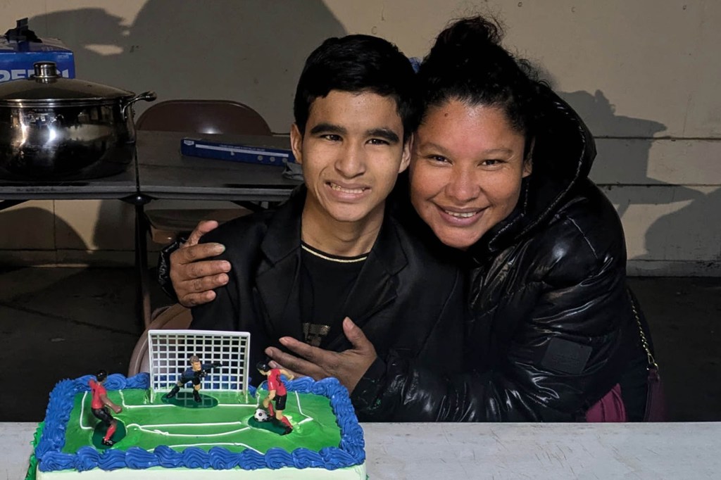 A smiling Emmanual beside his mother who has an arm around him. In front of them is a rectangular birthday cake decorated like a soccer field with small player figurines.