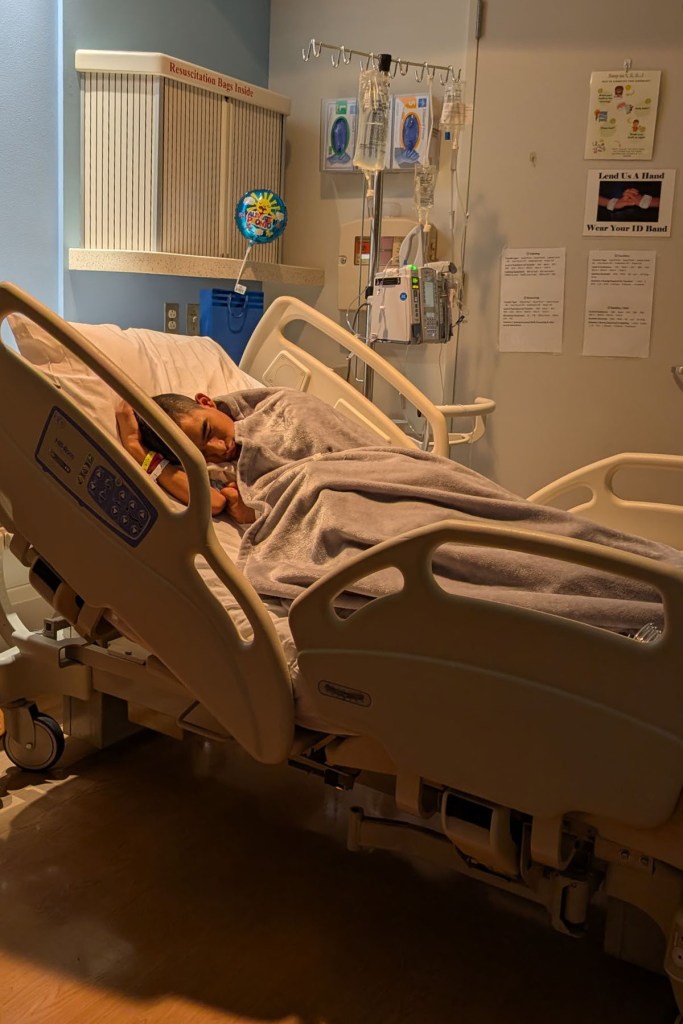 Emmanual lies on their side in a hospital bed covered with a gray blanket.