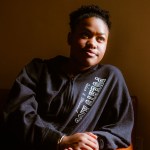 Adriana Del Orden sits on a couch in warm light, wearing a black hoodie with “Puerto Rico” written on it. She looks slightly upward, sunlight illuminating her face against a brown wall.
