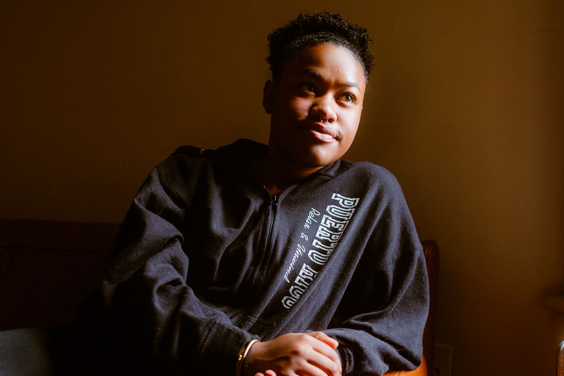 Adriana Del Orden sits on a couch in warm light, wearing a black hoodie with “Puerto Rico” written on it. She looks slightly upward, sunlight illuminating her face against a brown wall.