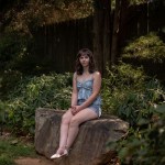 Ara Kareis sits on a large rock in a shaded garden, wearing a light blue sleeveless top and denim shorts, hands folded in her lap, looking calmly toward the camera.