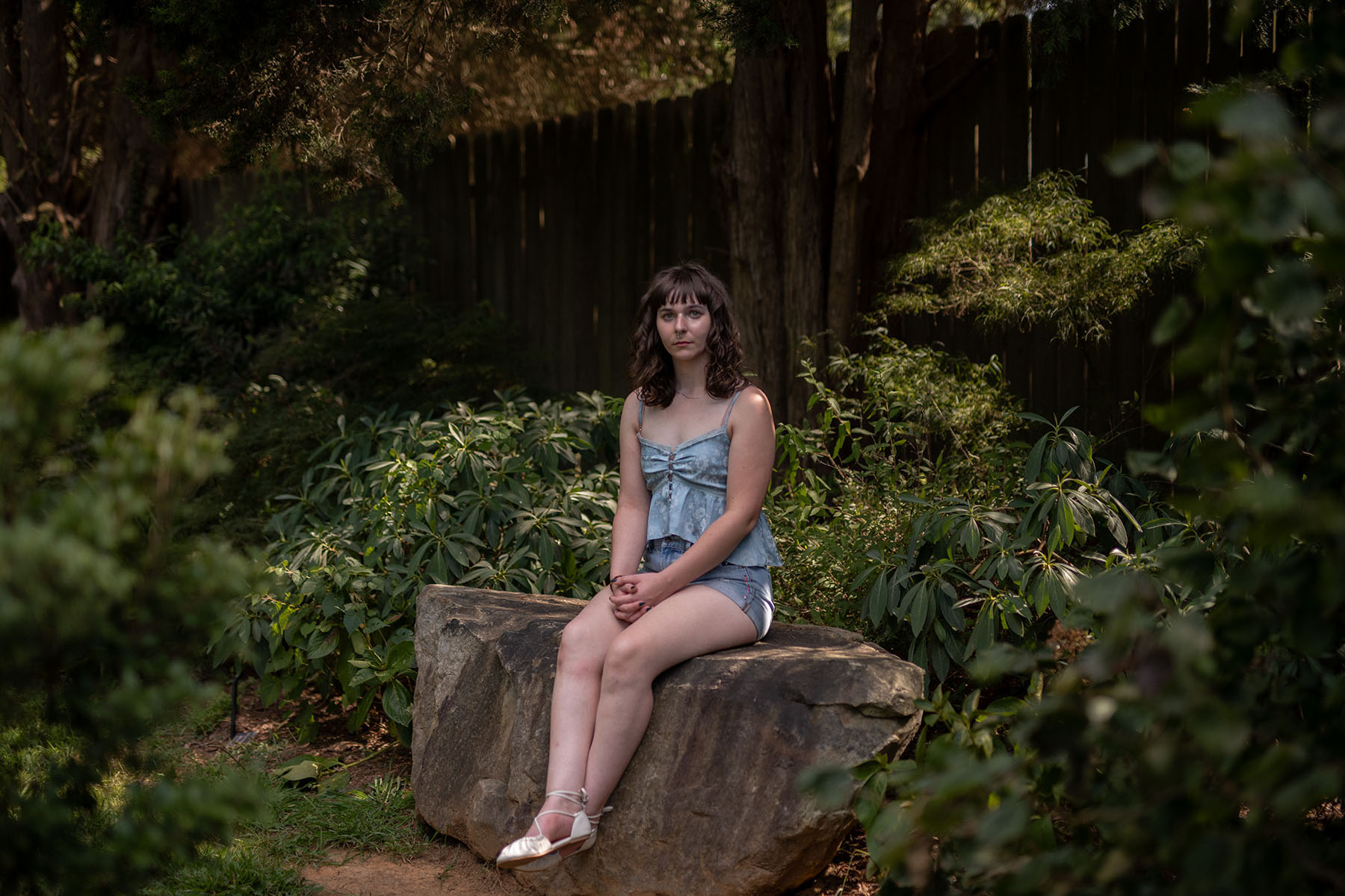 Ara Kareis sits on a large rock in a shaded garden, wearing a light blue sleeveless top and denim shorts, hands folded in her lap, looking calmly toward the camera.