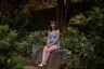 Ara Kareis sits on a large rock in a shaded garden, wearing a light blue sleeveless top and denim shorts, hands folded in her lap, looking calmly toward the camera.