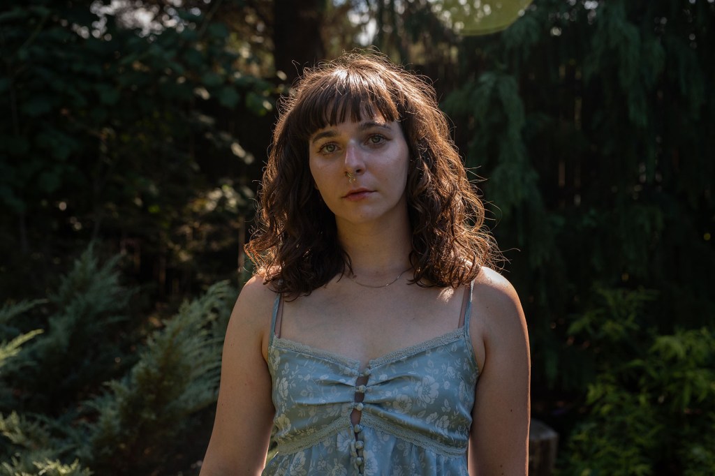 Ara Kareis stands outdoors in sunlight, looking directly at the camera. Her curly hair frames her face, and the light catches her shoulders against a backdrop of trees.