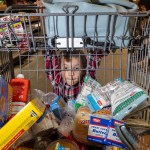 A young child looks through the bars of a grocery cart filled with food items. They hold onto the top of the cart while standing behind it in a supermarket aisle.