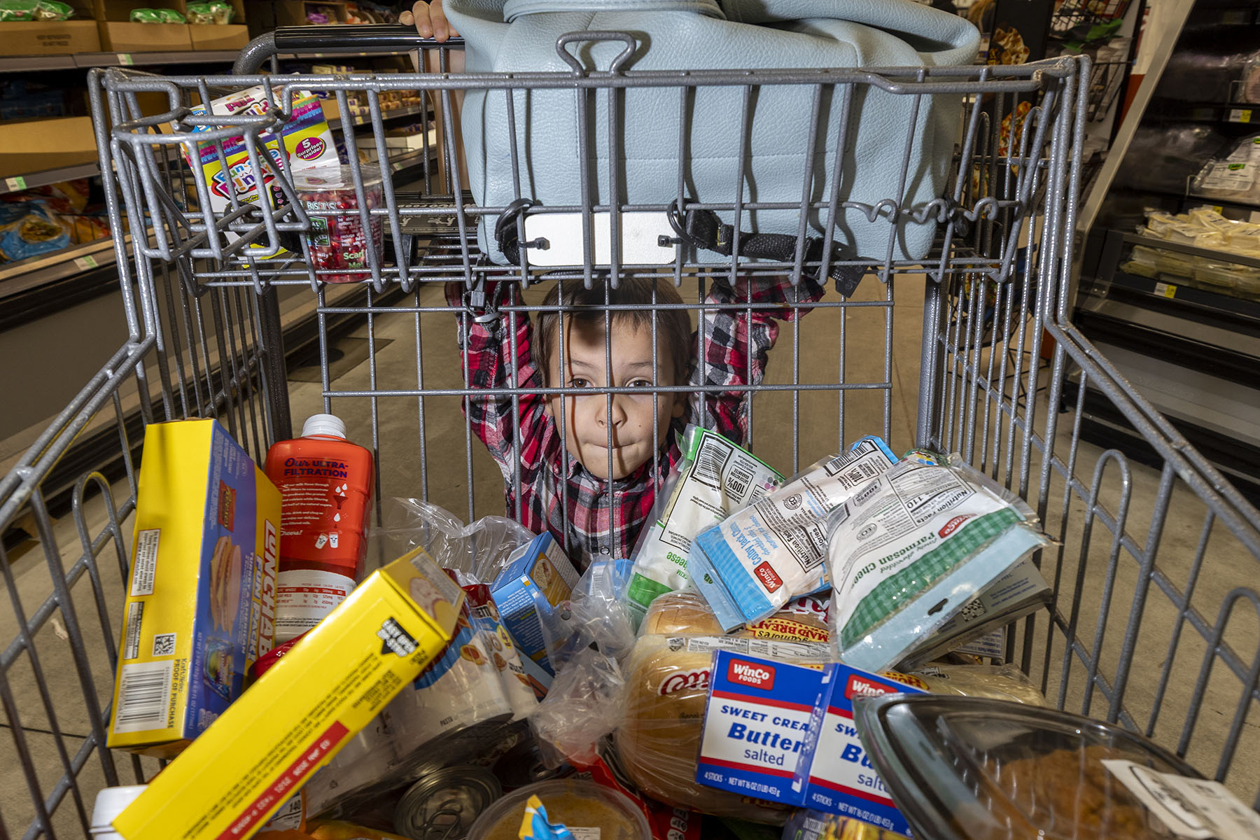 A young child looks through the bars of a grocery cart filled with food items. They hold onto the top of the cart while standing behind it in a supermarket aisle.